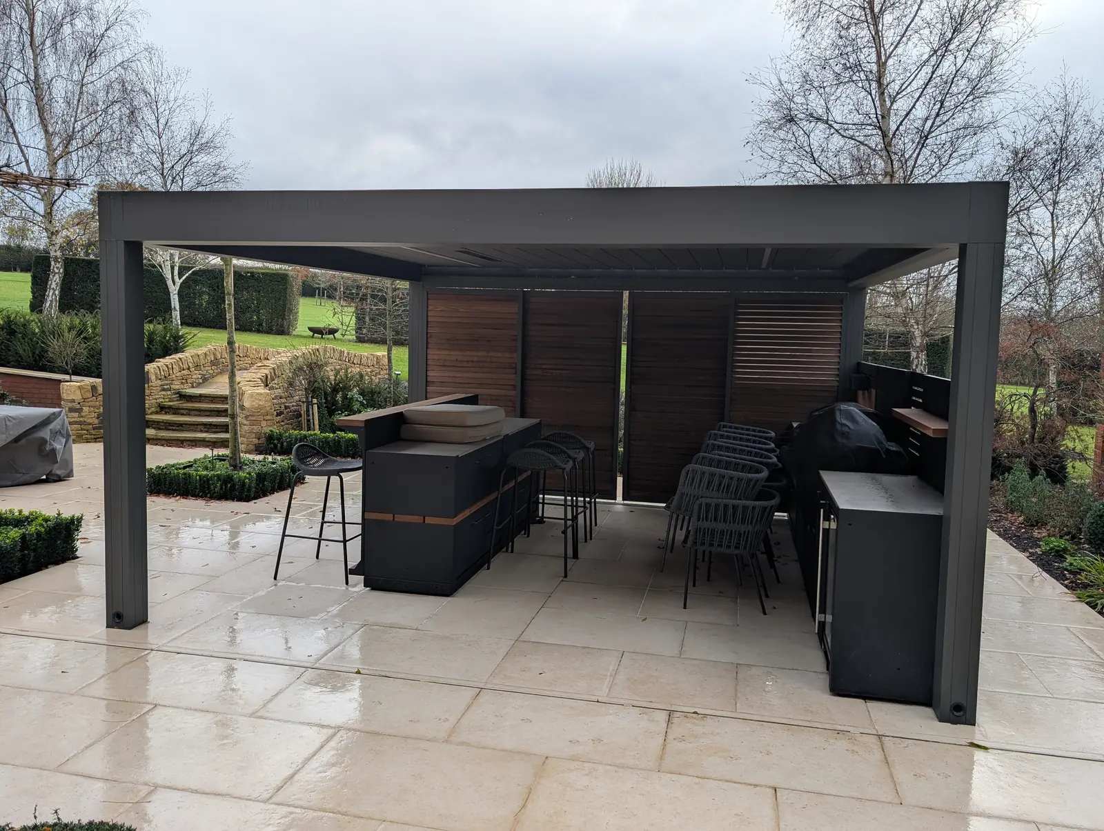 Anthracite aluminium pergola housing an outdoor kitchen and bar seating, with dry stone walls and garden steps beyond